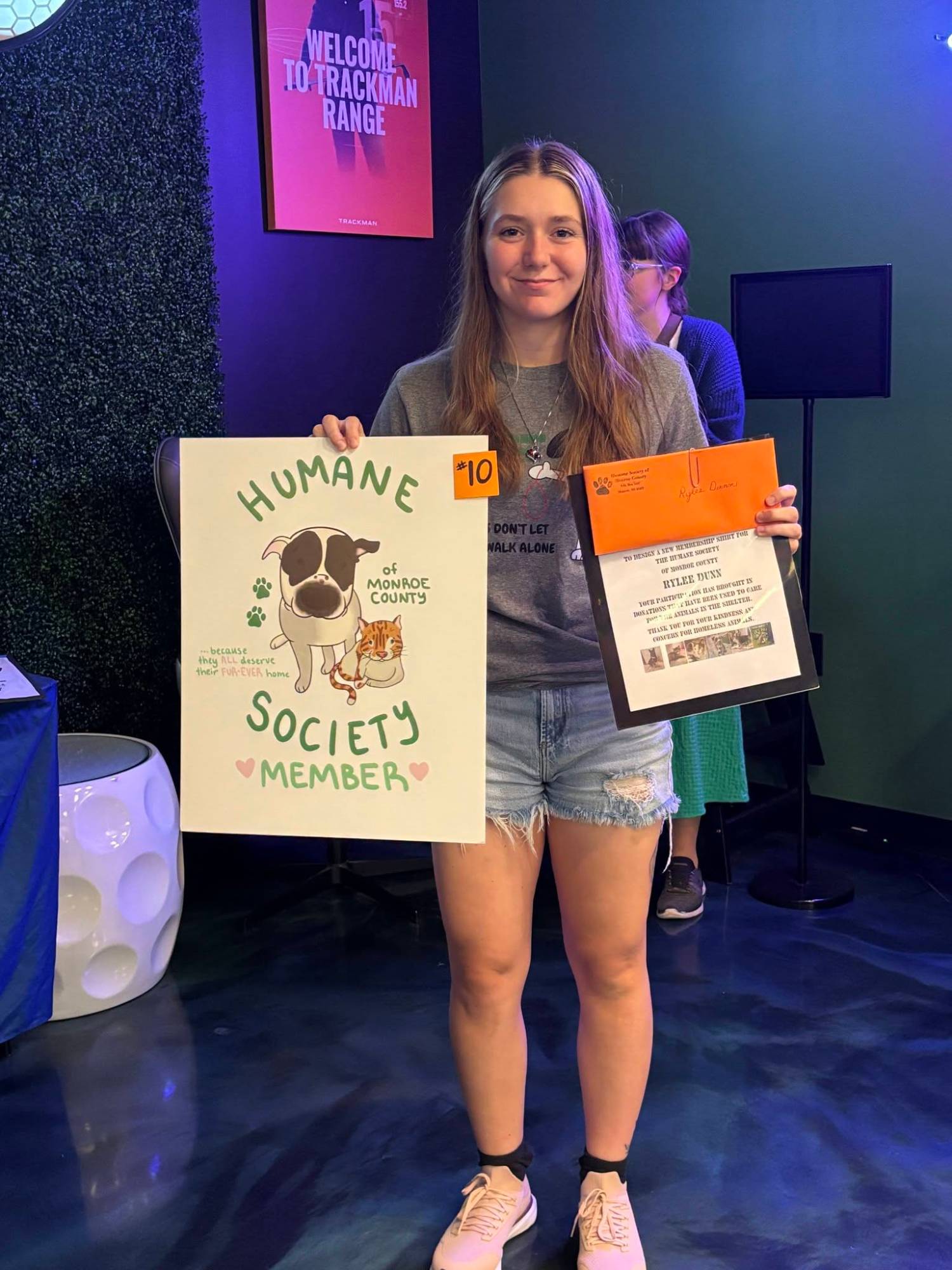 Rylee Dunn holding her second-place design, entry #10, reading Humane Society of Monroe County Member with an illustrated dog and cat.
